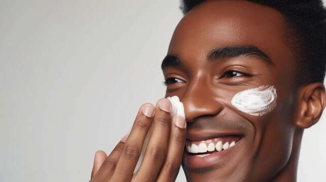 A young man with a radiant smile applying a white substance to his face possibly a skincare product or makeup against a neutral background.