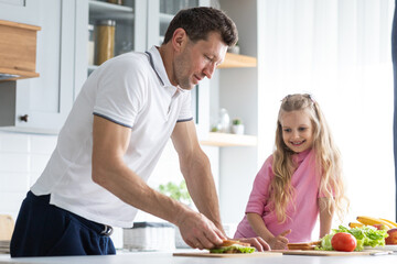 Obraz na płótnie Canvas A friendly family from father with daughter stand and manage in the kitchen. A young serious man dad gives his child culinary instructions to make sandwiches independently. The concept of mutual