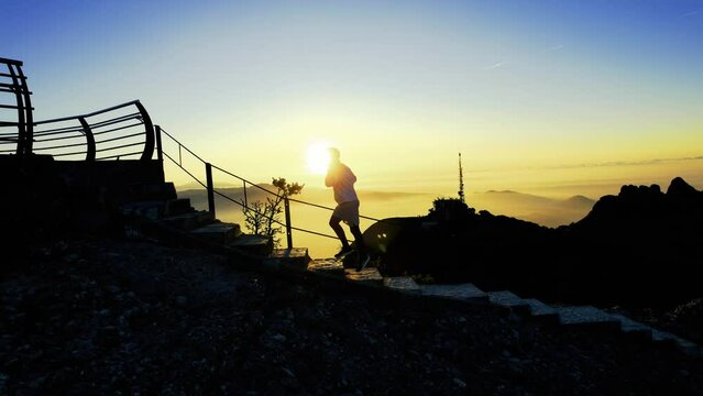 Silueta de deportista ascendiendo monta&ntilde;a con escalones al amanecer con la salida del sol detr&aacute;s por encima de las nubes