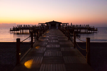 Fototapeta premium Early dark morning over sea pier is barely lit by the first rays of the sun and electric lamps.
