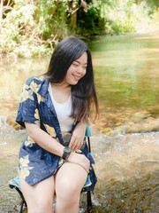 Asian woman enjoys sitting on a camping chair in a stream or a little waterfall. She smiles in a natural forest while on a camping trip with happiness.