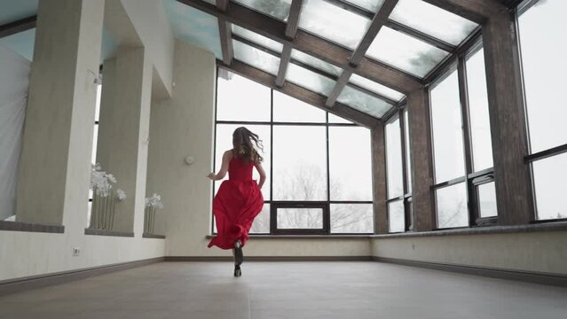 Woman in long red dress runs up to window. Young lady in art gallery orchestrates visually stunning episode in motion. Combination of elegance and glamour