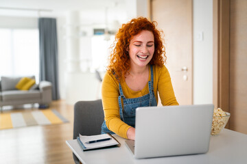 Young smiling woman typing on laptop in her home office. Redhead female with curly hair sitting in...