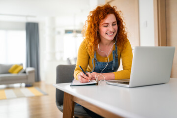 Young self-employed enthusiastic woman sitting in her home office and taking notes while having a joyful video call with her business partner.