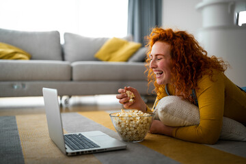 Beautiful young woman lying on the carpet and hugging a pillow, while watching comedy movie. Female...
