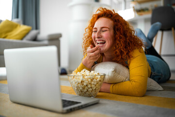 Attractive young woman, lying on the floor, hugging pillow and laughing at funny movie scene, while...