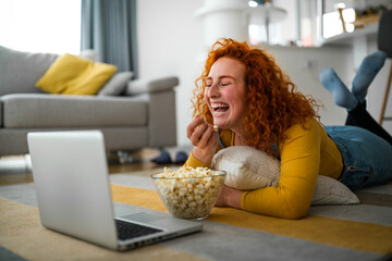 Young adult woman relaxing at home, watching comedy movies on laptop and eating popcorn in cozy,...