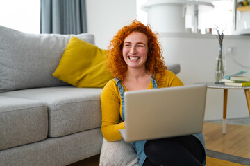 Young female college student sitting on the floor, holding a laptop in her lap, and smiling....