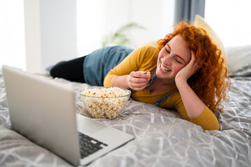 Young woman lying on bed in her bedroom, watching a movie on laptop and eating snack while laughing...