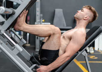 Young muscular man naked torso exercise using press machine to strength his legs in sport gym. Fit man training body workout doing heavy weights leg pushing equipment with quad muscles in fitness club