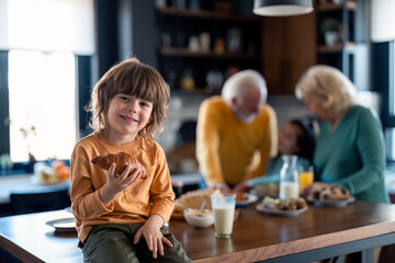 Portrait of a cute little boy having croissant during breakfast time in home atmosphere. He is sitting on the table in the kitchen and smiling at camera.