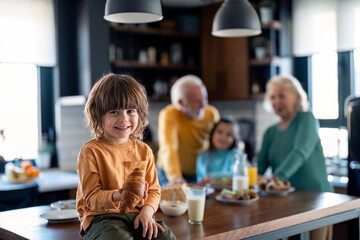 Portrait of a cute little boy having croissant during breakfast time at home. He is sitting on the table in the kitchen and smiling at camera.
