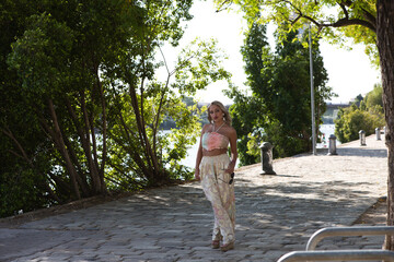 Young beautiful blonde woman dressed in embroidered trousers and crochet top by the river bank in seville. In the background green branches of a tree. Travel and holiday concept.