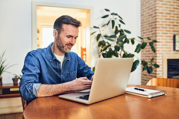 Smiling adult man using mobile phone and laptop sitting by table at home