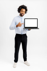 Smiling African-American businessman in formal wear showing laptop with empty display isolated on white. Male employee points with finger at blank screen, advertising new app, presentation of newelty