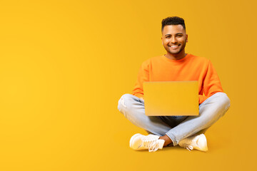 Smiling joyful indian man using laptop computer isolated on yellow. Portrait of male freelancer or student using computer sitting on the floor and looking at the camera