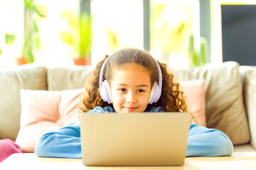 Happy schoolgirl doing homework at home. During pandemic or travel children continue learning process. Mixed-race KId receive assignments from teachers via laptop and headphones