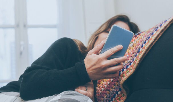 Relaxed Girl Using Smartphone Sitting On The Sofa At Home. Woman Chatting With Friends Sitting On A Couch In The Living Room With Warm Pajama In A Winter Morning. Leisure, Carefree And Relax Concept.