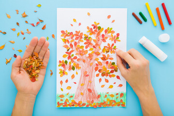 Woman hands gluing and creating tree shape from colorful leaves on white paper. Blue table...
