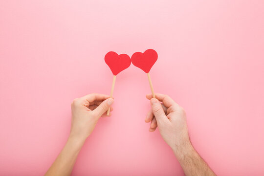Young adult woman and man hands holding red paper heart shapes on wooden sticks on light pink table background. Pastel color. Love concept. Closeup. Top down view.