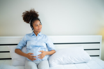 Young African American Pregnant woman sitting on bed listening music from headphone to relax, heal...