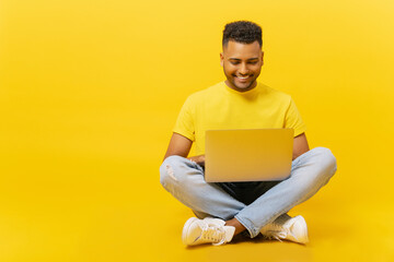Smiling indian man using laptop sitting on the floor in relaxed pose, isolated on yellow backdrop, carefree serene male student studying in the distance, freelancer working remotely