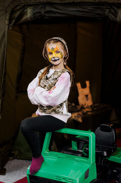 A girl, a child stands in military camouflage, a khaki uniform, a helmet with a painted face with strong tiger makeup near a children's car. Photography, Ukrainian pariot.