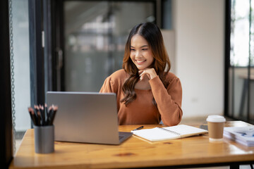Obraz premium Cheerful young woman in casual attire focusing on a laptop screen, working from a modern office.