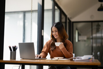 A young woman takes enjoy a hot beverage, with a smile, at her sunny office desk.