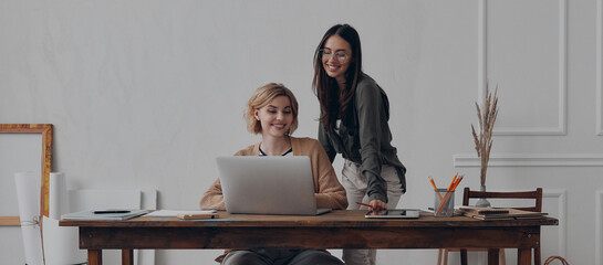 Two happy young businesswomen using laptop while working together at the creative office