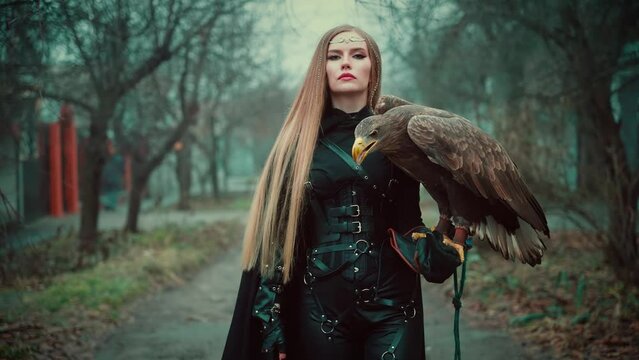 art portrait fantasy woman warrior holding white-tailed eagle wild bird on hand. Princess girl long blond hair. Lady queen assassin hunter black dress armor walking along street dark trees fog. 4k