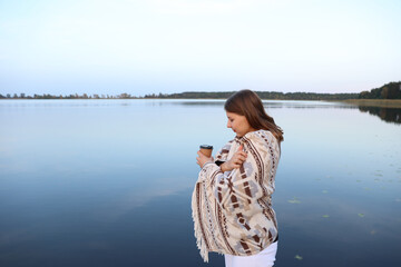 the girl is standing on a wooden pier near the lake wrapped in a blanket and drinking coffee....