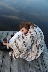 Fototapeta na wymiar the girl is sitting on a wooden pier near the lake wrapped in a blanket and drinking coffee. aesthetic photo. autumn vibe