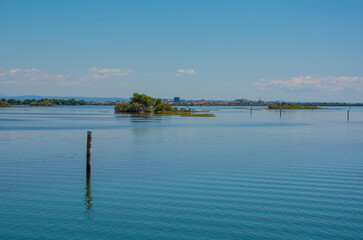 An island in the Grado section of the Marano and Grado Lagoon in Friuli-Venezia Giulia, NE Italy. Channel markers shows the edge of a navigable channel. Grado town is in background. August - obrazy, fototapety, plakaty