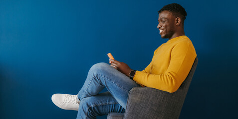 Happy young African man in casual wear using smart phone while sitting on the chair against the...