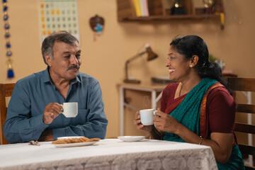 Joyful indian senior couples enjoying morning tea or coffee while spending together at home - concept of morning rituals or routines and refreshment or family bonding.