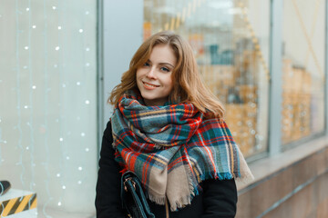 Happy beautiful young girl model with a sweet smile in a fashion vintage checkered scarf in a black coat with a handbag walks in the city with lights