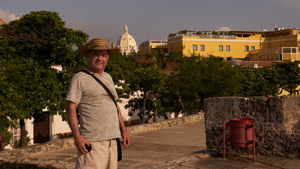Medium shot of an older man, dressed in casual clothing and wearing a 'vueltiao' hat, looking at the camera, with buildings and the tower of a church in the background in the walled city of Cartagena