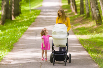 Happy smiling young adult mother and little daughter pushing white baby stroller and walking at...