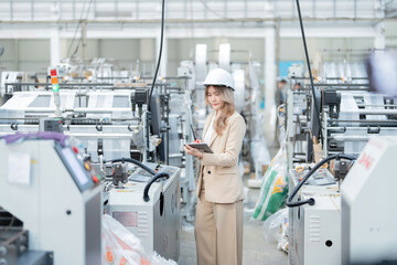 Asian female engineer manager Use a laptop to control machine systems in the plastics and steel production factory of a logistics business. Wear a suit, safety helmet, and have a radio.