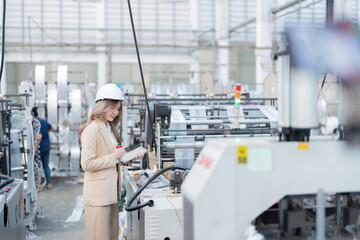 Work of Asian female engineer manager Standing at machinery and circuits in the plastics and steel manufacturing plant of a logistics business. Wearing a suit, safety helmet, and a laptop and a radio.