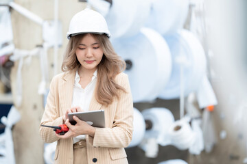 Caucasian female engineer with long hair holds a radio and uses a tablet to view circuits in an industrial factory at a plastic and steel production company. There are paper rolls in the warehouse.