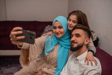 Two Arab parents and their daughter are sitting on the sofa while taking selfies together at home