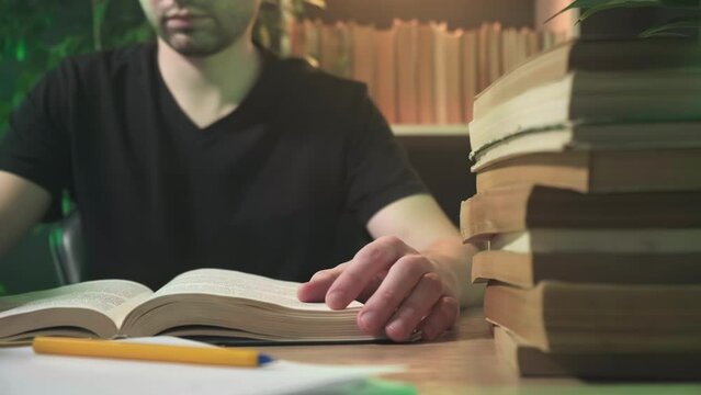 a man is reading a book in the library, a guy finishes reading a book and closes it, a man is reading a book at a table among other stacks of books
