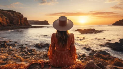 Foto op Canvas Young woman in hat sitting on the sand and looking at the sea at sunset. Solo travel, vacation concept. © Katerina Bond