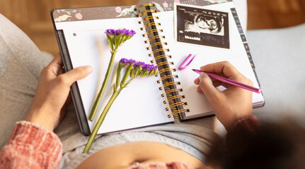 Pregnant woman writing in her diary looking at her first ultrasound in the living room. Woman...