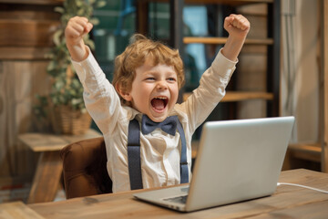 Portrait of a happy cheerful young business kid celebrating success with arms up in front of laptop