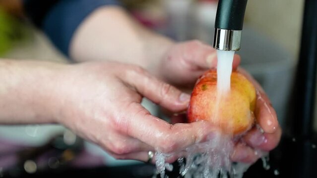 A man carefully washes an organic apple under the kitchen faucet. Hygiene, nitrates, cleanliness