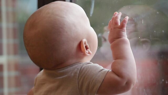 A child who is learning to stand licks a glass door with a blurred background in a beige jumpsuit