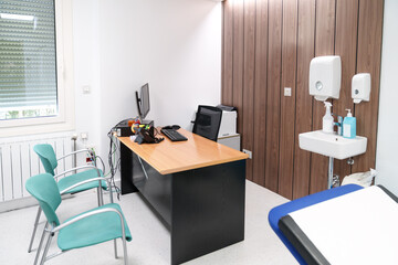 Clean and orderly doctor's consultation room with a wood desk, computer, hygiene amenities, and turquoise waiting chairs.